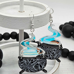 Könnte beinhalten: Ein Paar schwarze und weiße Kessel-Ohrringe mit blauen und silbernen Glitzerakzenten. Die Kessel haben ein Spinnennetz-Design und sind mit blauem und silbernem Glitzerrauch verziert.