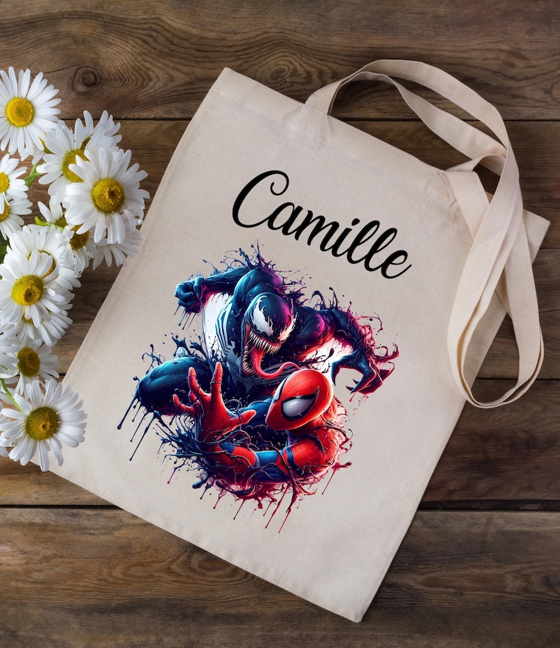 May include: A white canvas tote bag with a black and red graphic of Venom and Spider-Man fighting. The bag has a black handle and the name "Camille" printed in black cursive on the front.
