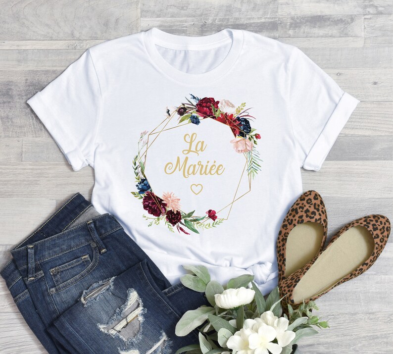 Puede incluir: Camiseta blanca con un dise&ntilde;o de corona floral y el texto dorado "La Mari&eacute;e". La corona presenta flores burdeos, rosas y azules. La camiseta se combina con vaqueros azules y bailarinas con estampado de leopardo.