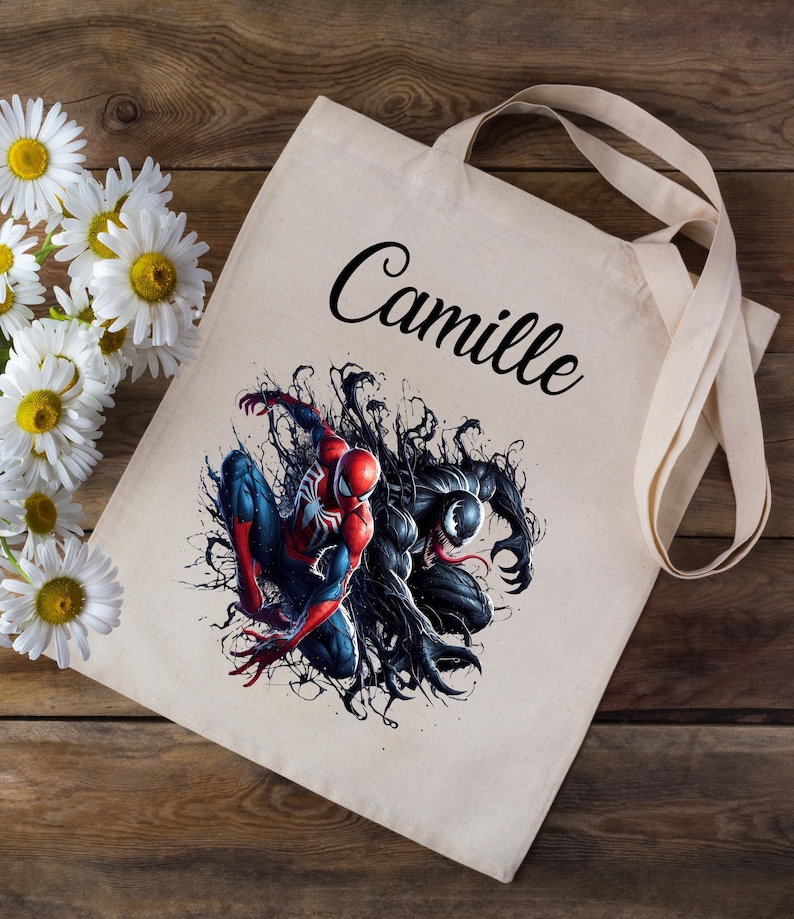 K&ouml;nnte beinhalten: Eine wei&szlig;e Canvas-Tragetasche mit schwarzen Henkeln. Die Tasche hat ein schwarzes und rotes Motiv von Spider-Man und Venom im Kampf. Der Name "Camille" ist in schwarzer Kursivschrift auf der Tasche aufgedruckt.