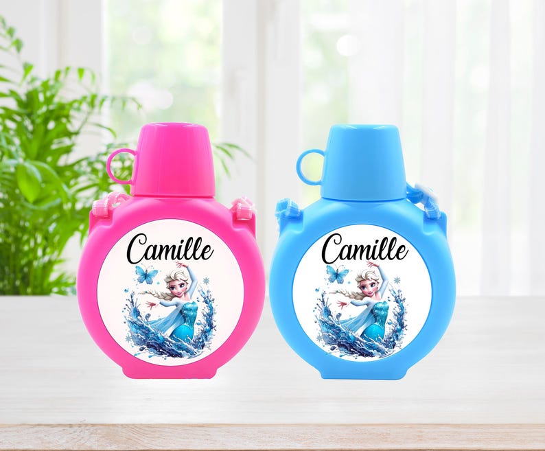 May include: Two round, pink and blue water bottles with matching caps. Each bottle features a white circular label with the name "Camille" and an image of a cartoon character in a blue dress. The bottles have a handle and a strap.