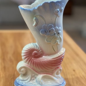 May include: A white ceramic vase with a pink seashell base and blue and white floral designs. The vase is shaped like a seashell and has a scalloped rim.