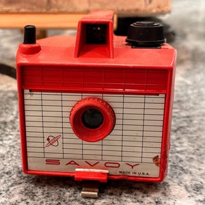 May include: A red Savoy box camera featuring a white front panel with a grid pattern, a central red lens, a black top knob, and a small black viewfinder. The front panel displays the text SAVOY and MADE IN U.S.A. with a small planet graphic.