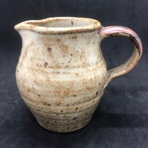 May include: A small, speckled, cream-coloured ceramic jug with a brown handle. The jug has a rounded body and a slightly flared lip.