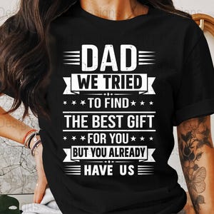 Pode incluir: T-shirt preto com um gráfico branco que diz "DAD WE TRIED TO FIND THE BEST GIFT FOR YOU BUT YOU ALREADY HAVE US." O design inclui estrelas e linhas horizontais.