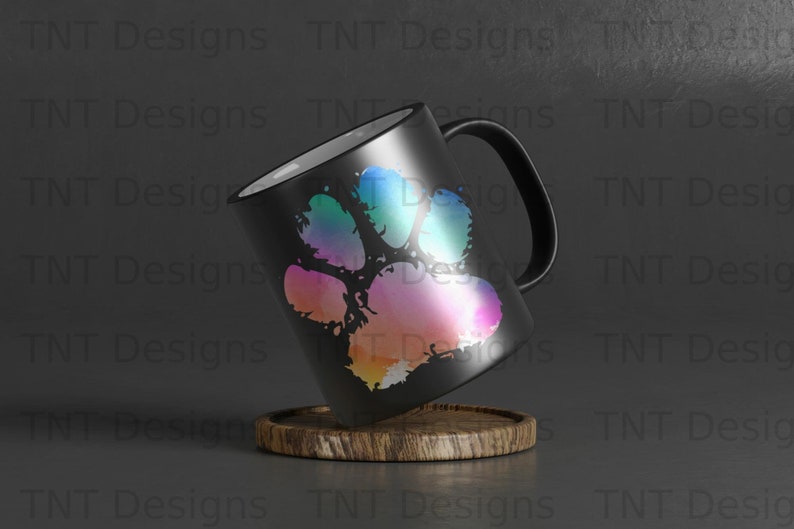 May include: A black ceramic mug with a colourful paw print design. The mug has a white interior and a handle. The mug is sitting on a wooden coaster.