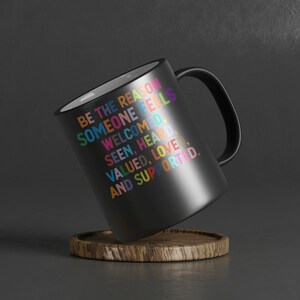 K&ouml;nnte beinhalten: Schwarze Kaffeetasse mit wei&szlig;em Innenraum und Henkel. Die Tasse tr&auml;gt eine bunte Botschaft mit dem Text: "Sei der Grund, warum sich jemand willkommen, gesehen, geh&ouml;rt, gesch&auml;tzt, geliebt und unterst&uuml;tzt f&uuml;hlt."