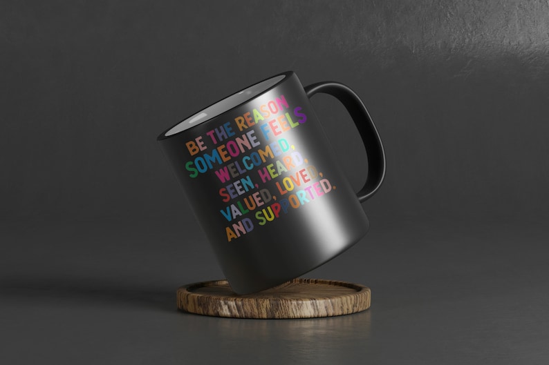 K&ouml;nnte beinhalten: Schwarze Kaffeetasse mit wei&szlig;em Innenraum und Henkel. Die Tasse tr&auml;gt eine bunte Botschaft mit dem Text: "Sei der Grund, warum sich jemand willkommen, gesehen, geh&ouml;rt, gesch&auml;tzt, geliebt und unterst&uuml;tzt f&uuml;hlt."