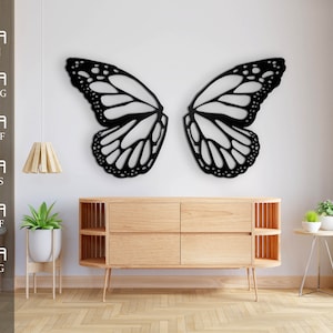 Könnte beinhalten: Schwarze Schmetterlings-Wanddekoration mit filigranen Flügeldetails. Der Schmetterling ist an einer weißen Wand montiert, im Vordergrund eine Kommode aus Holz und Topfpflanzen.