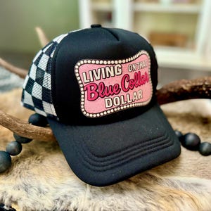 May include: Black trucker hat with a white and black chequered mesh back. The front of the hat features a pink and white sign that reads "Living on that Blue Collar Dollar."