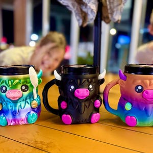 Könnte beinhalten: Drei bunte Tassen in Form von Highland-Kühen, jede mit einem Löffel. Die Tassen sind in Regenbogenfarben, Schwarz und Braun mit rosa Akzenten gehalten. Sie haben große Augen, Hörner und einen Henkel.