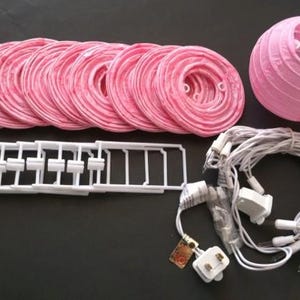 May include: A pink paper lantern kit with multiple round lanterns, a single assembled lantern, white plastic support structures, and a string of white lights. The kit is designed for decorative lighting.