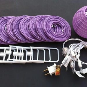 May include: A set of purple paper lanterns, string lights, and white plastic support structures. The lanterns are round and folded, with a matching round lantern assembled. The string lights have white wires and small bulbs.