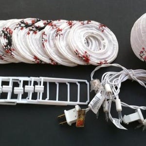 May include: A set of white paper lanterns with red floral designs, a white plastic frame, and a string of white lights. The lanterns are round and cylindrical, perfect for decorative lighting.