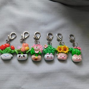 May include: A collection of six handmade keychains featuring miniature, smiling plant pot designs. Each keychain showcases a different plant, including red berries, tulips, and cacti, with adorable faces. The keychains have silver-colored clasps.
