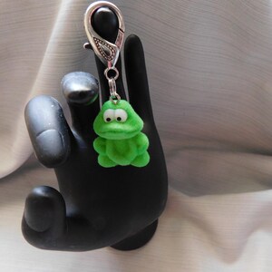 May include: A green frog keychain with a silver clasp. The frog has large eyes and a textured, fuzzy appearance. The keychain is attached to a silver metal clasp.