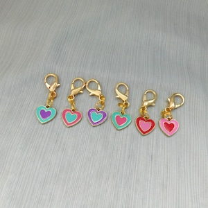 May include: Six gold heart-shaped stitch markers with different colored enamel. The hearts have a double layer with a contrasting color on the inside. The colors are purple, teal, pink, and red.