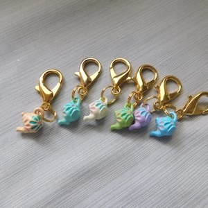 May include: Six colorful charm pendants with gold-colored lobster clasps. Each charm is a small, snail-shaped design in pastel colors: pink, blue, green, and purple. The charms are attached to the clasps with small gold-colored rings.
