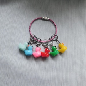 May include: A circular pink wire bracelet featuring six small, colorful rubber duck charms attached with silver-toned metal rings. The ducks are light blue, bright blue, pink, red, green, and yellow, each with a distinct orange beak.