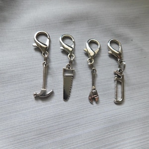 May include: Four silver-colored charms with lobster clasps. The charms are shaped like tools: a hammer, a saw, a wrench, and a pipe wrench. The charms are hanging against a gray background.