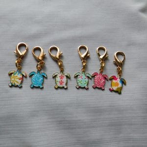 May include: Six colorful turtle charms with gold-colored lobster clasps. The turtles have various patterns, including floral and geometric designs, in shades of blue, green, pink, and yellow. The charms are displayed on a gray fabric surface.