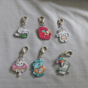 May include: Six colorful cat-themed keychains with silver metal rings. The keychains feature a cat sleeping on a book, a cat sleeping in a red armchair, a cat holding a smaller cat, a cat floating in a pink donut, a cat in a suitcase, and a cat sitting in a blue beach chair under an umbrella.