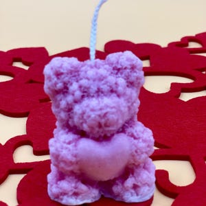 May include: A pink teddy bear candle holding a heart in its paws. The candle is on a red background with heart cutouts.