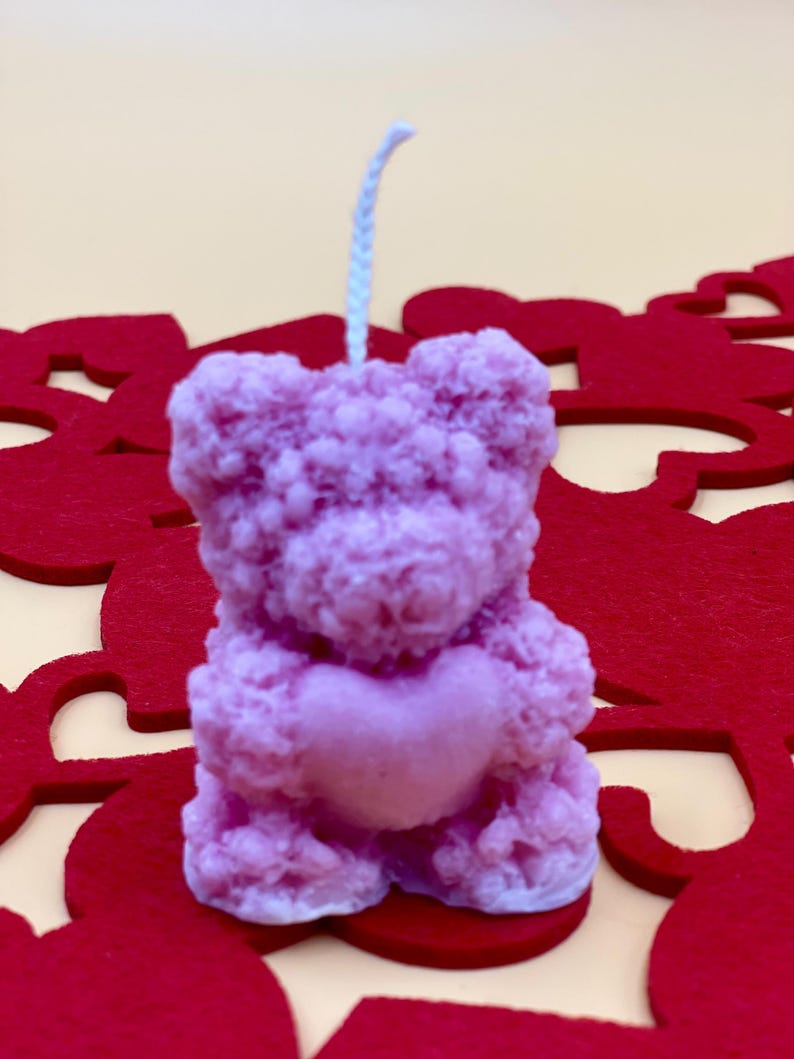 May include: A pink teddy bear candle holding a heart in its paws. The candle is on a red background with heart cutouts.