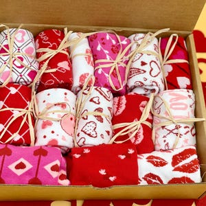 May include: A box filled with pairs of colorful socks with heart and love themed patterns. The socks are tied with twine and arranged in a neat pattern.
