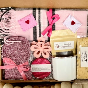 May include: A pink and black plaid blanket, a pair of pink fuzzy socks, a pink flower-shaped hair clip, a white mug, a bag of coffee beans, a pink bath bomb, and a package of caramel vanilla biscotti in a brown gift box.