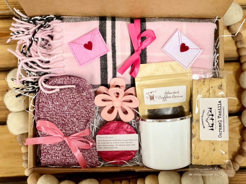 May include: A pink and black plaid blanket, a pair of pink fuzzy socks, a pink flower-shaped hair clip, a white mug, a bag of coffee beans, a pink bath bomb, and a package of caramel vanilla biscotti in a brown gift box.