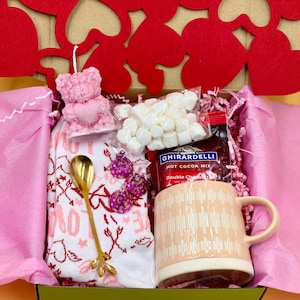May include: A pink gift box filled with Valentine's Day themed items, including a pink bear candle, a mug, a bag of marshmallows, a bag of Ghirardelli hot cocoa mix, a gold spoon, and a pink and white patterned towel.