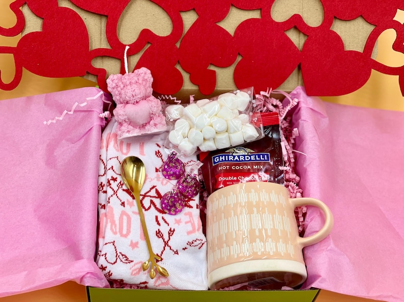 May include: A pink gift box filled with Valentine's Day themed items, including a pink bear candle, a mug, a bag of marshmallows, a bag of Ghirardelli hot cocoa mix, a gold spoon, and a pink and white patterned towel.