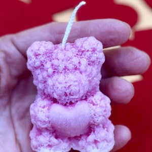 May include: A pink bear-shaped candle with a heart in its paws. The candle has a textured surface and a white wick.