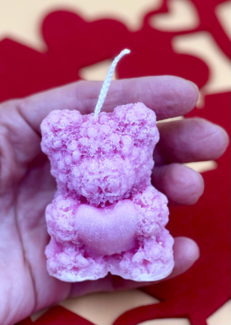 May include: A pink bear-shaped candle with a heart in its paws. The candle has a textured surface and a white wick.