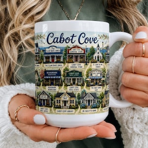 Op de afbeelding: Een witte keramische mok met een kleurrijke geïllustreerde kaart van Cabot Cove. Het ontwerp bevat diverse gebouwen met labels zoals The Clam Bake Diner, Cabot Cove General Store, Bookmark Bookstore en Cabot Cove Library.