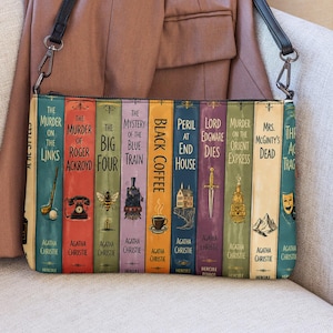May include: A rectangular crossbody bag featuring a graphic print of multiple Agatha Christie book spines, including titles like The Murder of Roger Ackroyd and Black Coffee, with a black shoulder strap and a mauve jacket draped behind it.