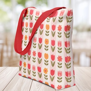 May include: A floral tote bag with a red strap. The bag features a repeating pattern of tulips and flowers in shades of red, orange, and pink on a cream background with vertical stripes. The bag is sitting on a wooden surface.