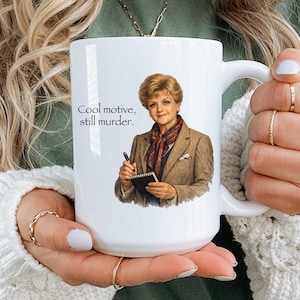 Op de afbeelding: Een witte keramische mok met een grafische illustratie van een persoon met kort blond haar, gekleed in een beige blazer en een sjaal met patroon, die een pen en notitieblok vasthoudt. De tekst "Cool motive, still murder." staat in het zwart gedrukt.