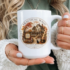 May include: A white ceramic mug featuring a circular graphic with a house, autumn trees, a black cat, and a cauldron. The text reads Practical Magic, 1995, The Owens Sisters, and Witches Welcome Salem, Massachusetts in a decorative circular border.