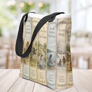 May include: A white tote bag with black fabric handles featuring a printed design of multiple book spines from the Little House series by Laura Ingalls Wilder, including titles like On the Banks of Plum Creek and By the Shores of Silver Lake.