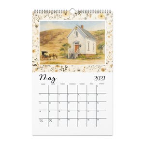 May include: A wall calendar for May 2021 featuring a watercolor illustration of a white schoolhouse with a brown door and a horse-drawn carriage on a dirt path. The border is decorated with a floral pattern in muted yellow, brown, and green tones.