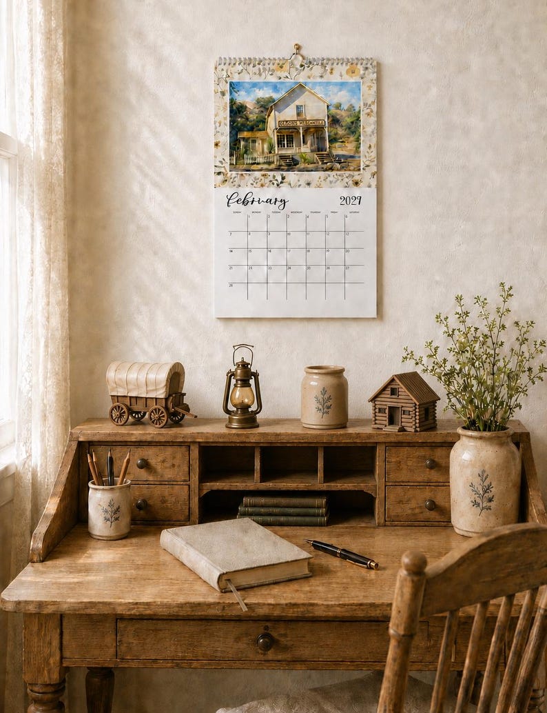 May include: A wall calendar for February 2027 featuring a watercolor illustration of a rustic building labeled 'General Merchandise'. The calendar is displayed above a wooden desk with a miniature wagon, a lantern, a ceramic jar, and a log cabin model.