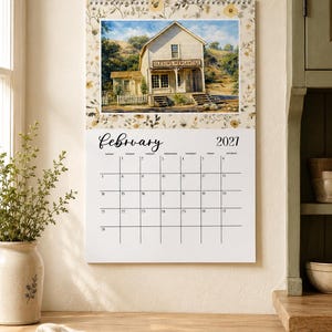 May include: A wall calendar for February 2027 featuring a top image of a wooden building labeled Oleson's Mercantile. The calendar has a floral border pattern in shades of yellow and brown, with a white grid for dates and black text.