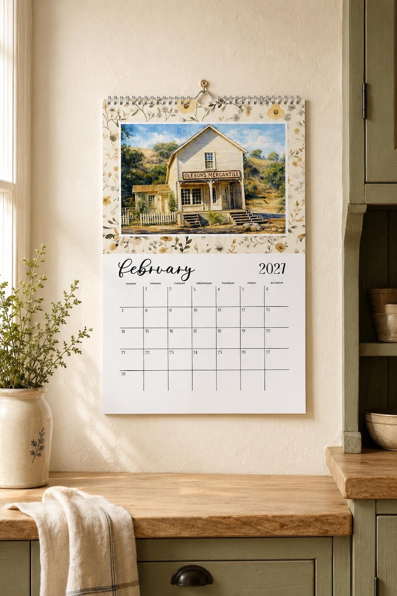 May include: A wall calendar for February 2027 featuring a top image of a wooden building labeled Oleson's Mercantile. The calendar has a floral border pattern in shades of yellow and brown, with a white grid for dates and black text.