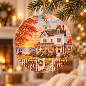 May include: A round ceramic ornament featuring a detailed illustration of a white Victorian house with a tower, surrounded by autumn foliage, a white picket fence, and two figures standing by a lake at sunset with a small sailboat.