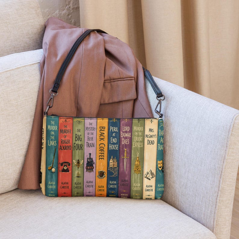 May include: A rectangular crossbody bag featuring a graphic print of multiple Agatha Christie book spines, including titles like The Murder of Roger Ackroyd and Black Coffee, with a black shoulder strap and a mauve jacket draped behind it.