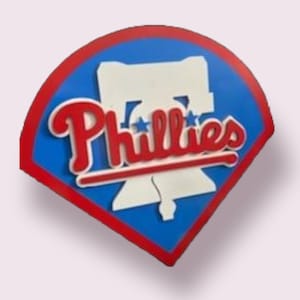 May include: A baseball-shaped emblem featuring the Philadelphia Phillies logo. The design includes a red border, a blue background, and the team name in red script. The Liberty Bell is also visible.