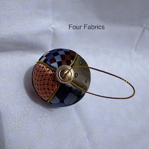 Op de afbeelding: Een ronde, veelkleurige stoffen ornament met een gouden lus en een gouden knoop. De ornament is gemaakt van vier verschillende stoffen: een bruin en wit geruit, een blauw en wit gestreept, een bruin en oranje geweven en een groen en bruin herringbone stof. De ornament is op een witte achtergrond met een sneeuwvlokkenpatroon.