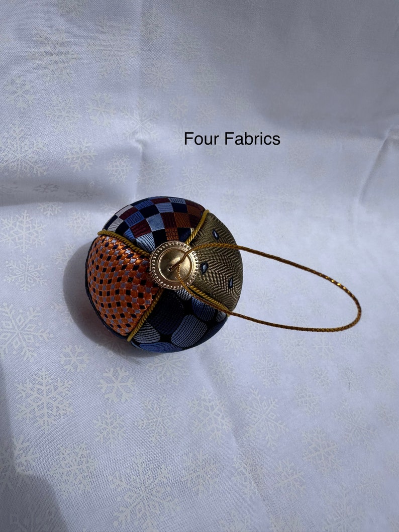 Op de afbeelding: Een ronde, veelkleurige stoffen ornament met een gouden lus en een gouden knoop. De ornament is gemaakt van vier verschillende stoffen: een bruin en wit geruit, een blauw en wit gestreept, een bruin en oranje geweven en een groen en bruin herringbone stof. De ornament is op een witte achtergrond met een sneeuwvlokkenpatroon.